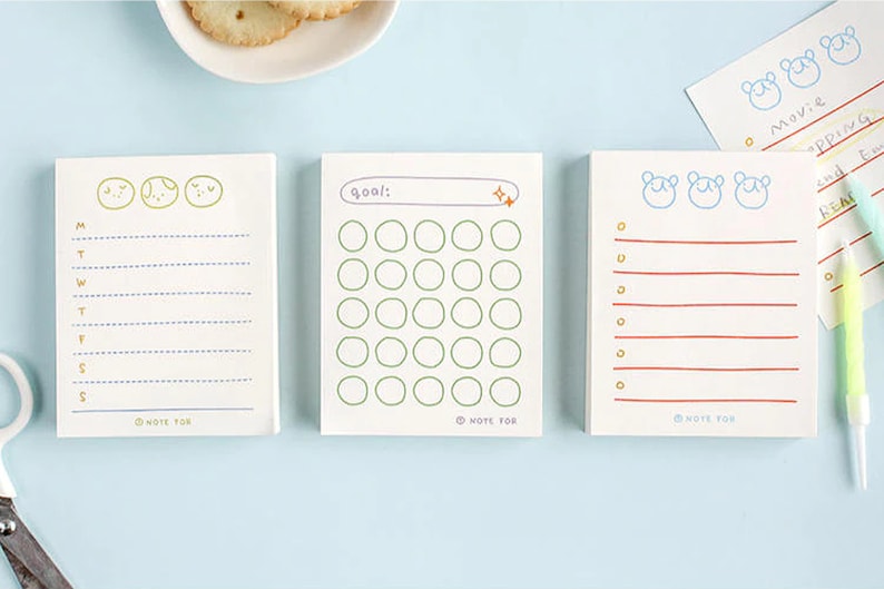 May include: Three white notepads with different designs. The first notepad has a weekly planner with a blue and gold design. The second notepad has a goal tracker with a green and orange design. The third notepad has a lined notepad with a red and gold design.