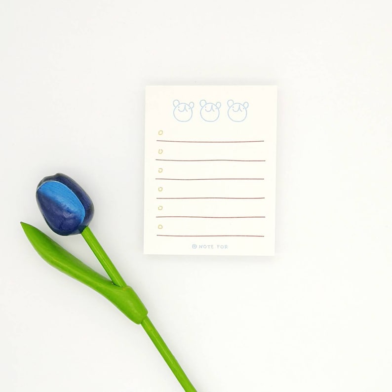 May include: A white notepad with a blue line design and three bear faces at the top. The notepad has a brown line at the bottom with the text "@NOTE FOR". A blue and green tulip is in the lower left corner of the image.
