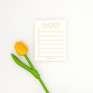 May include: A white weekly planner notepad with a cute cartoon design of three smiling faces. The notepad has a lined section for each day of the week, Monday through Sunday. The notepad is on a white background with a yellow and green tulip.