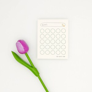 May include: A white notepad with a goal tracker. The notepad has a line for writing a goal and 36 circles to track progress. The notepad has a gold star sticker on the top right corner. A pink tulip is in the lower left corner of the image.