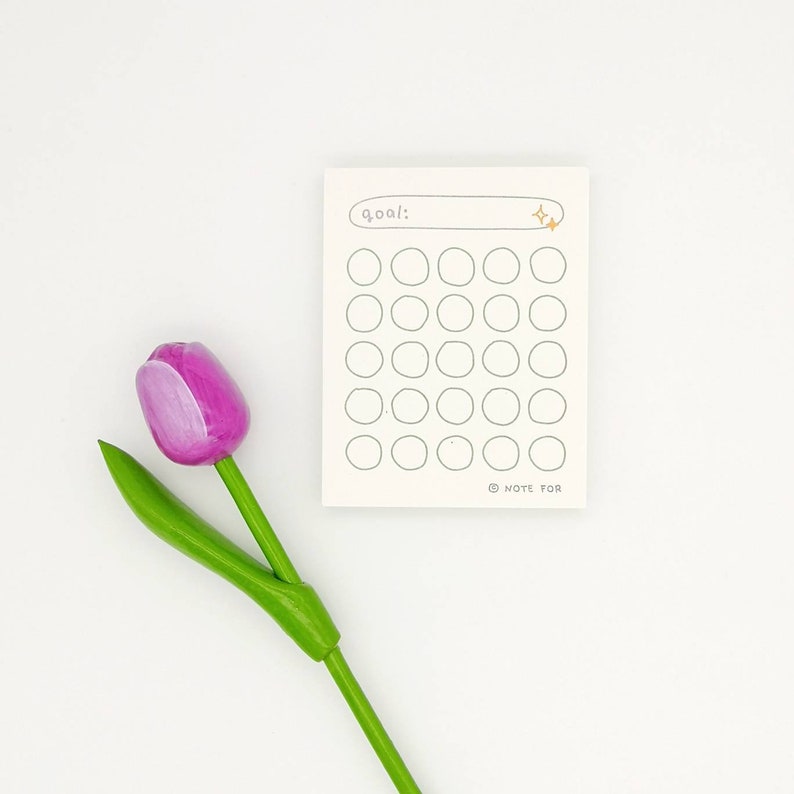 May include: A white notepad with a goal tracker. The notepad has a line for writing a goal and 36 circles to track progress. The notepad has a gold star sticker on the top right corner. A pink tulip is in the lower left corner of the image.