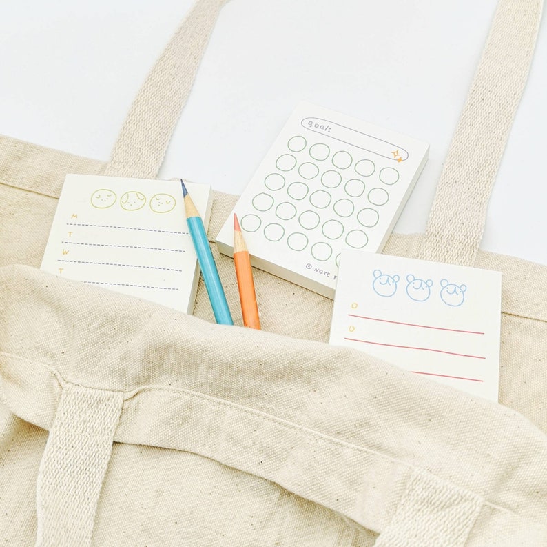 May include: A beige canvas tote bag with three different notepads inside. The notepads have different designs, including a checklist, a weekly planner, and a notepad with cute cartoon faces. Two colored pencils are also inside the bag.