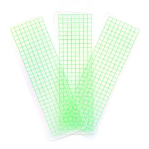 May include: Three clear plastic rulers with a green grid pattern. The rulers are stacked on top of each other.