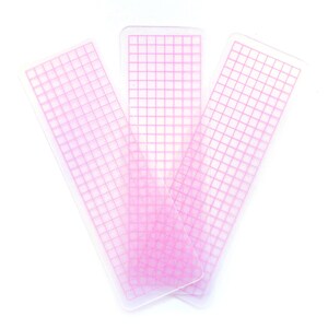 May include: Three pink gridded plastic rulers, each with a different orientation, on a white background.