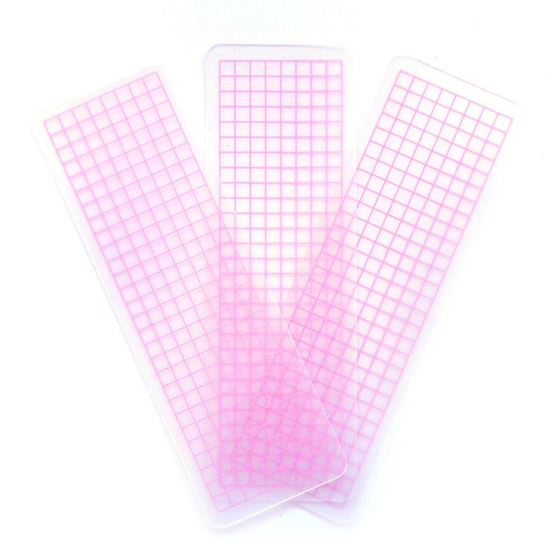 May include: Three pink gridded plastic rulers, each with a different orientation, on a white background.