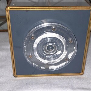 Op de afbeelding: Een vierkante, grijs en goud omlijste decoratieve doos met een heldere, cirkelvormige, meerlagige glazen inzet. De glazen inzet heeft een reeks kleine, zilverkleurige klinknagels. Op de achtergrond is een geometrisch, goud omlijst object zichtbaar.