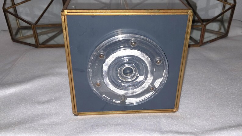 Op de afbeelding: Een vierkante, grijs en goud omlijste decoratieve doos met een heldere, cirkelvormige, meerlagige glazen inzet. De glazen inzet heeft een reeks kleine, zilverkleurige klinknagels. Op de achtergrond is een geometrisch, goud omlijst object zichtbaar.