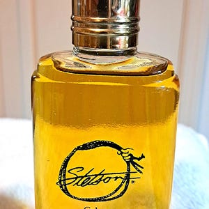May include: A rectangular glass cologne bottle with a gold-colored cap. The bottle contains a yellow liquid and features the "Stetson" logo in a black circular design. The label reads "Cologne" and "3.0 FL. OZ."
