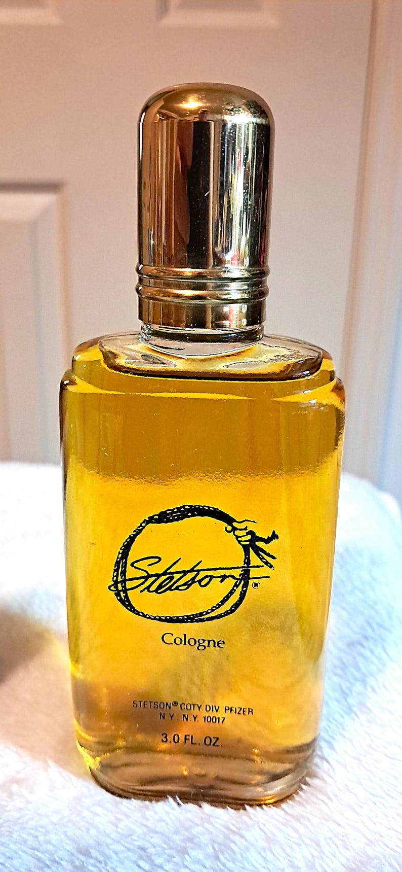 May include: A rectangular glass cologne bottle with a gold-colored cap. The bottle contains a yellow liquid and features the "Stetson" logo in a black circular design. The label reads "Cologne" and "3.0 FL. OZ."