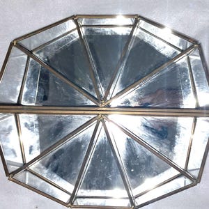 Op de afbeelding: Een geometrische decoratieve doos van glas en metaal met een scharnierend deksel. De doos bestaat uit meerdere driehoekige en trapeziumvormige glazen panelen, omlijst door dun, goudkleurig metaal. De doos is open en onthult het interieur.