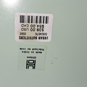 Op de afbeelding: Twee witte stickers met zwarte tekst en een streepjescode liggen op een lichtgroen oppervlak. De bovenste sticker leest "URBAN OUTFITTERS" en bevat de tekst "$39.00 USD" en "$54.00 CAD". De onderste sticker zegt "Made in India Fabriqu&eacute; en Inde" en "UD Home".