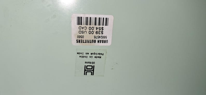 Op de afbeelding: Twee witte stickers met zwarte tekst en een streepjescode liggen op een lichtgroen oppervlak. De bovenste sticker leest "URBAN OUTFITTERS" en bevat de tekst "$39.00 USD" en "$54.00 CAD". De onderste sticker zegt "Made in India Fabriqu&eacute; en Inde" en "UD Home".
