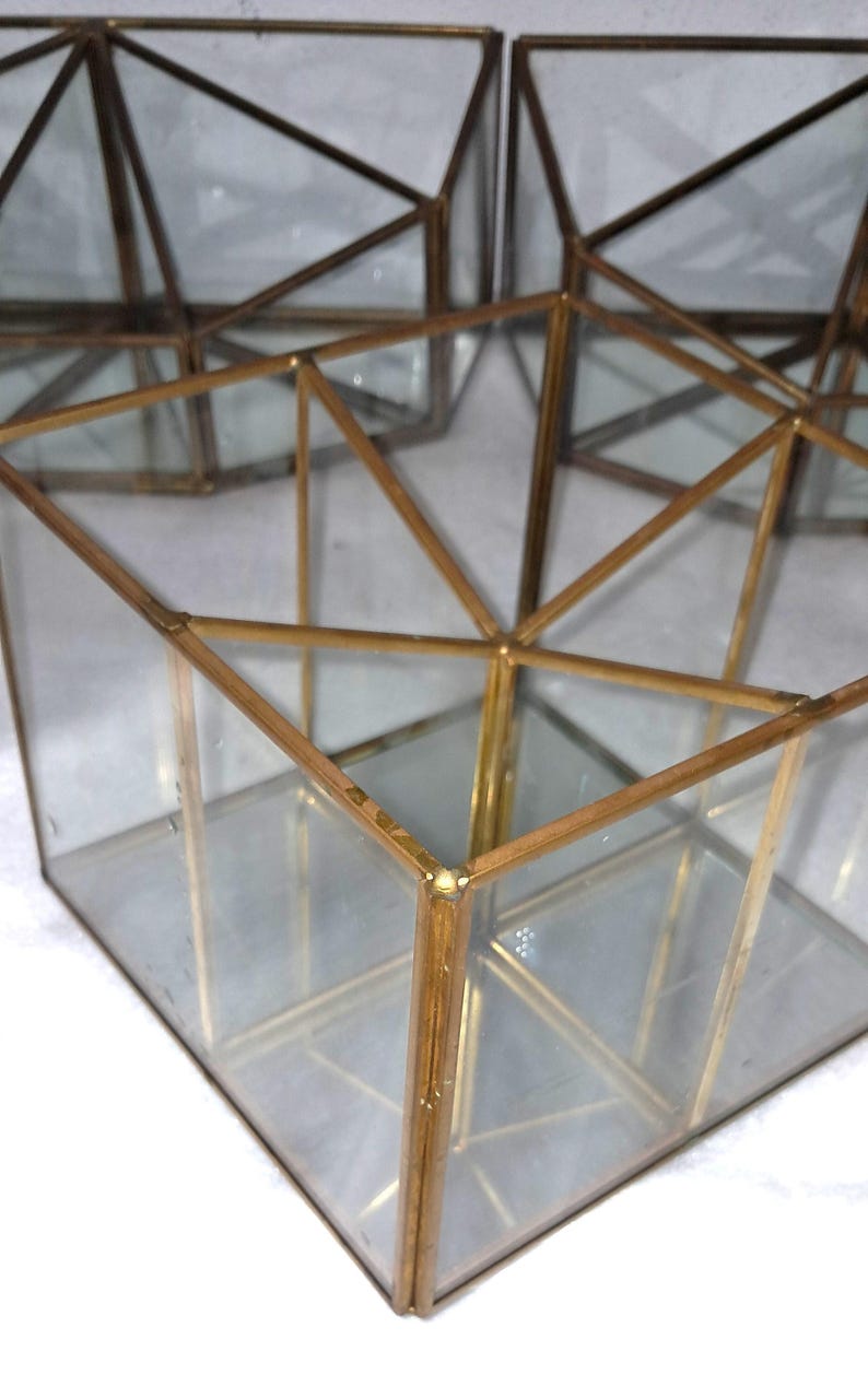 Op de afbeelding: Geometrisch terrarium van glas en messing. De heldere glazen panelen worden omlijst door dun, goudkleurig metaal. Het terrarium heeft een kubusvorm met driehoekige accenten. Er zijn meerdere terrariums zichtbaar, met een witte achtergrond.