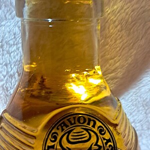 May include: A vintage Avon Wild Country cologne bottle. The bottle is a clear, amber-colored glass with a silver-colored cap. The bottle has a circular emblem with a bull's head and the words "Avon Wild Country".