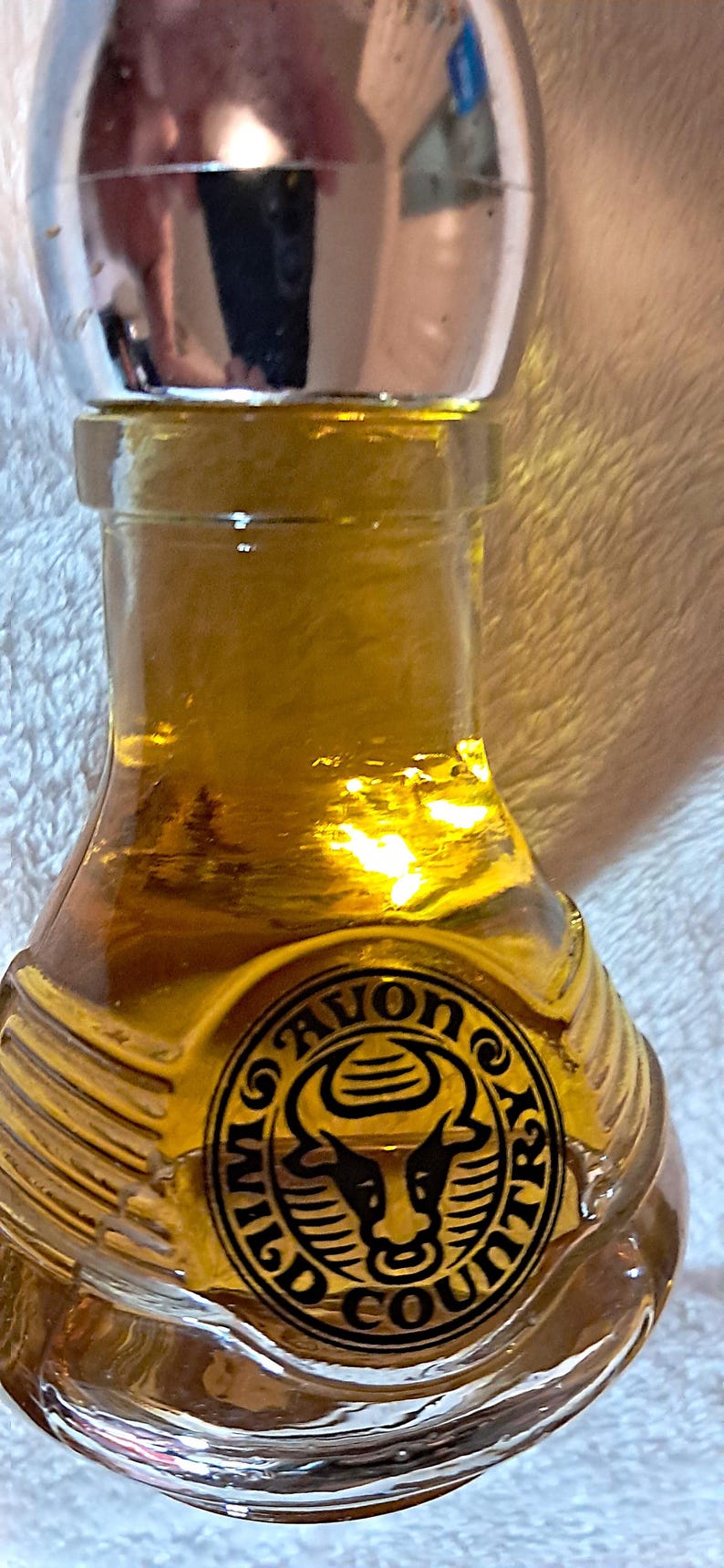 May include: A vintage Avon Wild Country cologne bottle. The bottle is a clear, amber-colored glass with a silver-colored cap. The bottle has a circular emblem with a bull's head and the words "Avon Wild Country".