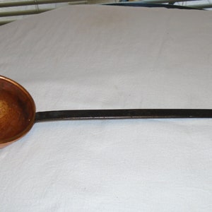 May include: A copper ladle with a long, black metal handle. The ladle has a rounded bowl and a small loop at the end of the handle.
