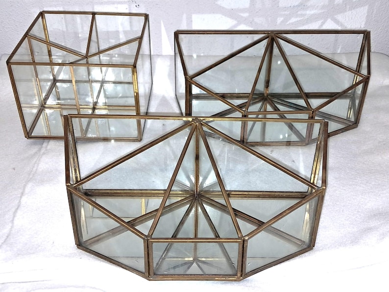 Op de afbeelding: Drie geometrische glazen terrariums met messingkleurige metalen frames. De terrariums hebben verschillende vormen, waaronder een kubus, een rechthoekig prisma en een vijfhoekige vorm. Het glas is helder en de frames zijn warm goudkleurig.