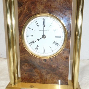 May include: A Howard Miller mantel clock with a gold-tone frame and a burl wood face. The clock face is white with black Roman numerals and hands. The clock is rectangular and sits on a gold base.