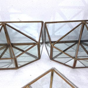 Op de afbeelding: Geometrische terrariums met heldere glazen panelen en bronskleurige metalen frames. De terrariums hebben een driehoekig ontwerp en zijn leeg. De afbeelding toont drie terrariums, waarvan er twee naast elkaar staan.