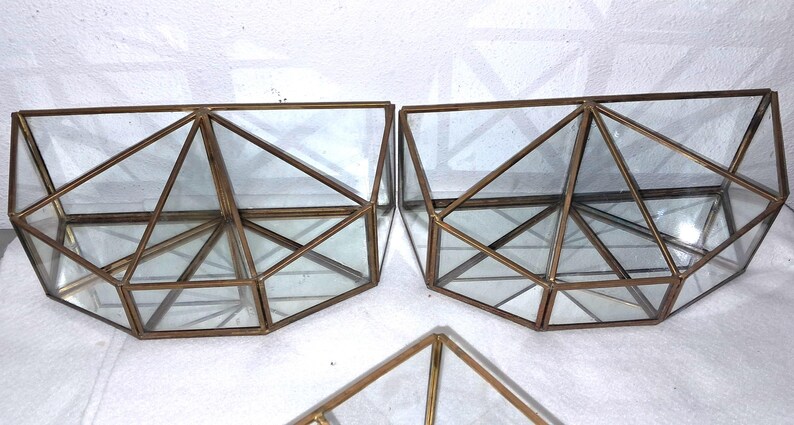 Op de afbeelding: Geometrische terrariums met heldere glazen panelen en bronskleurige metalen frames. De terrariums hebben een driehoekig ontwerp en zijn leeg. De afbeelding toont drie terrariums, waarvan er twee naast elkaar staan.