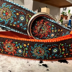 May include: A brown dog collar with a colorful sunburst pattern and orange trim. The collar has a silver metal buckle.