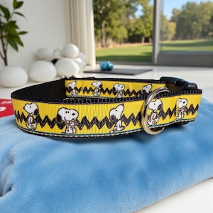 May include: A yellow dog collar featuring a repeating pattern of Snoopy characters and black zigzags. The collar has a black backing, a silver metal ring, and a black plastic buckle. The collar is resting on a light blue surface.