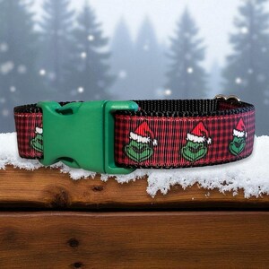 May include: A pet collar featuring a red and black buffalo plaid pattern with a repeating graphic of a green character wearing a red Santa hat. The collar includes a solid green plastic side-release buckle and a metal D-ring for attachment.