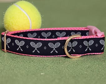 May include: A navy blue dog collar with a pink trim and a gold-colored metal buckle. The collar features a pattern of white tennis rackets. A yellow tennis ball rests in the background.