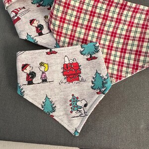 以下が含まれることがあります： 3つの犬用お祭りバンダナ。2つは、ピーナッツのキャラクターとクリスマスツリーが描かれたグレーです。3つ目は赤と白のチェック柄です。バンダナは布製で、折りたたまれています。