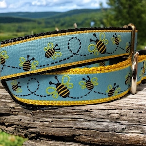 May include: A blue and yellow dog collar with a repeating pattern of bees and a black border. The collar has a silver D-ring.