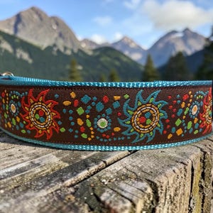 May include: A brown pet collar featuring a vibrant embroidered pattern of stylized suns and small geometric shapes in red, turquoise, yellow, and green. The collar is finished with bright turquoise trim along the top and bottom edges.