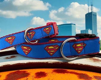 May include: Two blue pet collars featuring a repeating Superman logo pattern in red and yellow. Each collar includes a silver metal D-ring for leash attachment and a plastic side-release buckle, with red fabric trim along the edges of the straps.