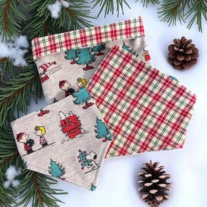 以下が含まれることがあります： クリスマスをテーマにした3つの布製品。1つは、赤と緑のチェック柄のトリムと、漫画のキャラクターとクリスマスツリーが描かれたベージュの背景を持つ長方形の容器です。他の2つは正方形で、1つはチェック柄、もう1つは漫画のキャラクターです。