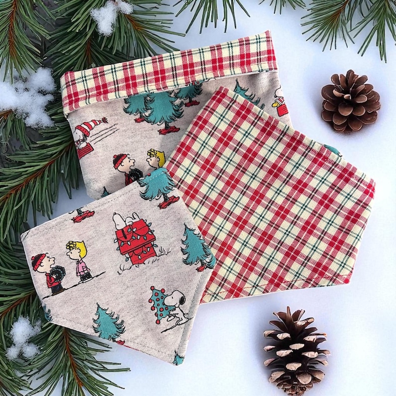 以下が含まれることがあります： クリスマスをテーマにした3つの布製品。1つは、赤と緑のチェック柄のトリムと、漫画のキャラクターとクリスマスツリーが描かれたベージュの背景を持つ長方形の容器です。他の2つは正方形で、1つはチェック柄、もう1つは漫画のキャラクターです。