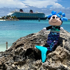 Puede incluir: Un juguete de peluche de sirena azul y morado con una corona de flores azules se sienta en una roca con un gran crucero en el fondo.