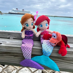 Puede incluir: Dos peluches de sirena, uno con escamas moradas y doradas y el otro con escamas azules y doradas, sentados en una superficie de madera con un juguete de peluche de cangrejo rojo. Las sirenas tienen detalles de flores rosas y moradas en el pelo.