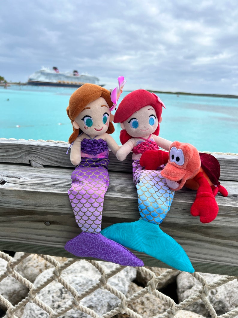 Puede incluir: Dos peluches de sirena, uno con escamas moradas y doradas y el otro con escamas azules y doradas, sentados en una superficie de madera con un juguete de peluche de cangrejo rojo. Las sirenas tienen detalles de flores rosas y moradas en el pelo.