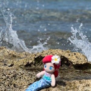 Puede incluir: Una mu&ntilde;eca de sirena de peluche con cabello rojo, una flor blanca y una cola azul y verde se sienta en una superficie rocosa cerca del oc&eacute;ano.