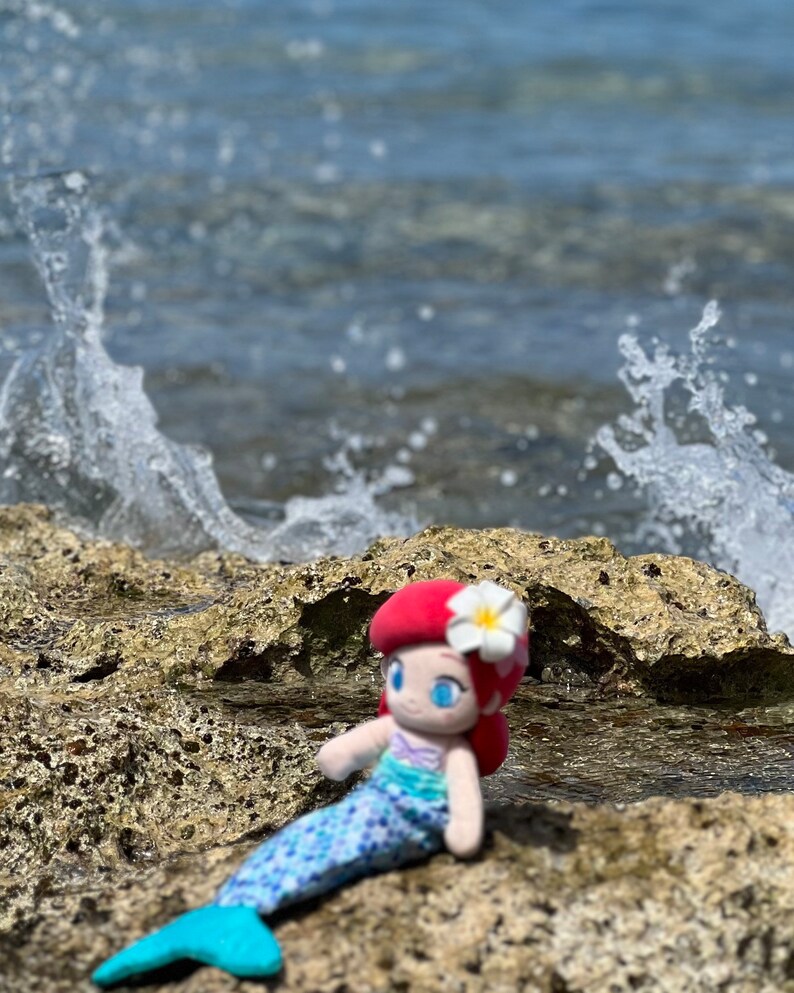 Puede incluir: Una mu&ntilde;eca de sirena de peluche con cabello rojo, una flor blanca y una cola azul y verde se sienta en una superficie rocosa cerca del oc&eacute;ano.