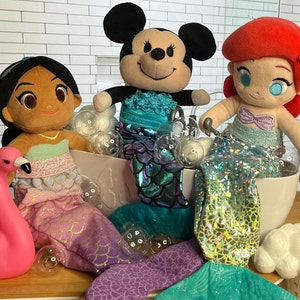 Puede incluir: Tres mu&ntilde;ecas de peluche vestidas de sirenas en una ba&ntilde;era llena de burbujas. Las mu&ntilde;ecas son una mu&ntilde;eca de piel morena con cabello negro, una mu&ntilde;eca de cabello rojo con una corona y una mu&ntilde;eca rat&oacute;n blanco y negro. Las mu&ntilde;ecas llevan todas colas de sirena y tienen accesorios de conchas.