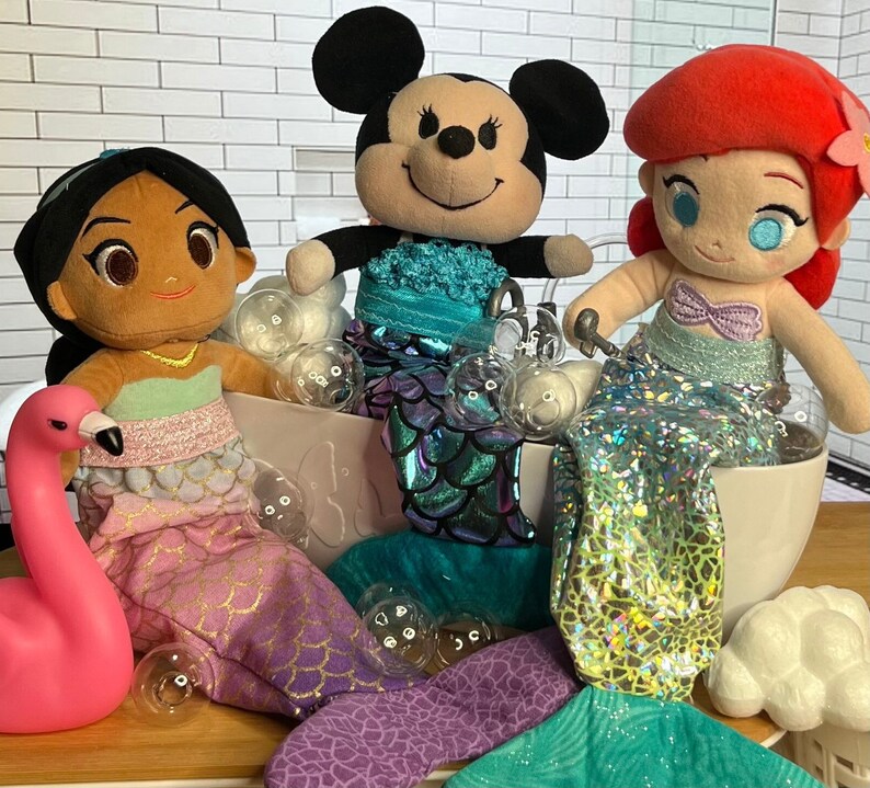 Puede incluir: Tres mu&ntilde;ecas de peluche vestidas de sirenas en una ba&ntilde;era llena de burbujas. Las mu&ntilde;ecas son una mu&ntilde;eca de piel morena con cabello negro, una mu&ntilde;eca de cabello rojo con una corona y una mu&ntilde;eca rat&oacute;n blanco y negro. Las mu&ntilde;ecas llevan todas colas de sirena y tienen accesorios de conchas.
