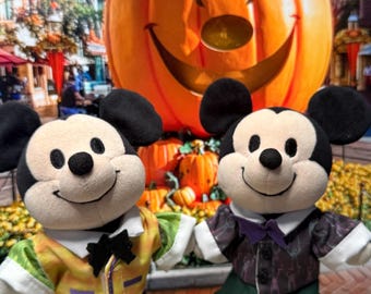 May include: Two plush Mickey Mouse figures dressed in Halloween-themed outfits. One wears a yellow vest, purple pants, and light green clogs with the text BOO. The other wears a dark patterned vest, green pants, and black clogs with ghost charms.