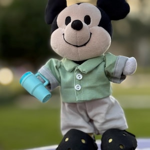 May include: A plush Mickey Mouse doll wearing a green button-down shirt, khaki pants, and black shoes. The doll is holding a light blue tumbler. The background is blurred green.