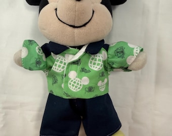 Könnte beinhalten: Eine Mickey-Maus-Plüschfigur mit cremefarbenem Gesicht, schwarzen Ohren und gelben Füßen. Die Figur trägt ein grünes Hemd mit Kragen und einem weißen, sich wiederholenden Muster aus Mickey-Maus-Köpfen und Globen sowie eine marineblaue Hose.