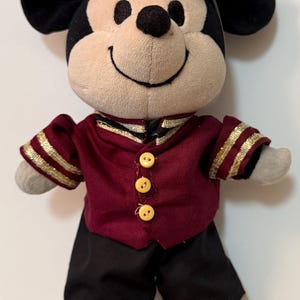 May include: A plush Mickey Mouse doll wearing a maroon and gold uniform with black pants and a matching hat. The doll has a smiling face, black ears, and yellow feet. The uniform has gold trim and three yellow buttons.