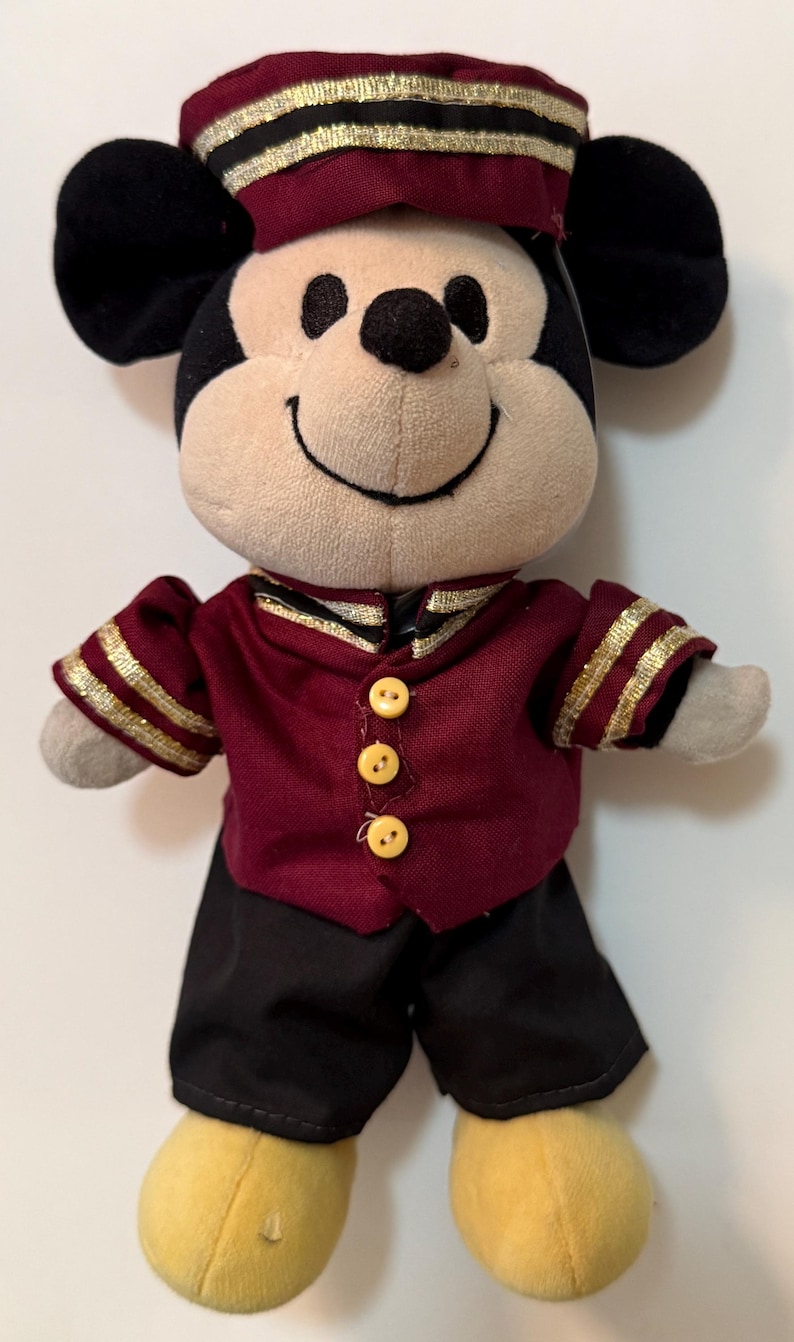 May include: A plush Mickey Mouse doll wearing a maroon and gold uniform with black pants and a matching hat. The doll has a smiling face, black ears, and yellow feet. The uniform has gold trim and three yellow buttons.