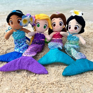 Puede incluir: Cuatro mu&ntilde;ecas de sirena de peluche con colas y tops de diferentes colores. Las mu&ntilde;ecas est&aacute;n sentadas en una playa de arena.
