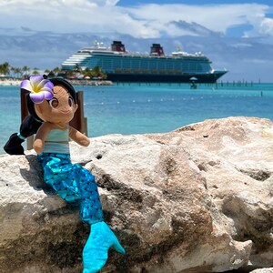 Puede incluir: Una mu&ntilde;eca de sirena de peluche con cabello casta&ntilde;o, una flor morada y una cola azul se sienta en una roca con un gran crucero en el fondo.