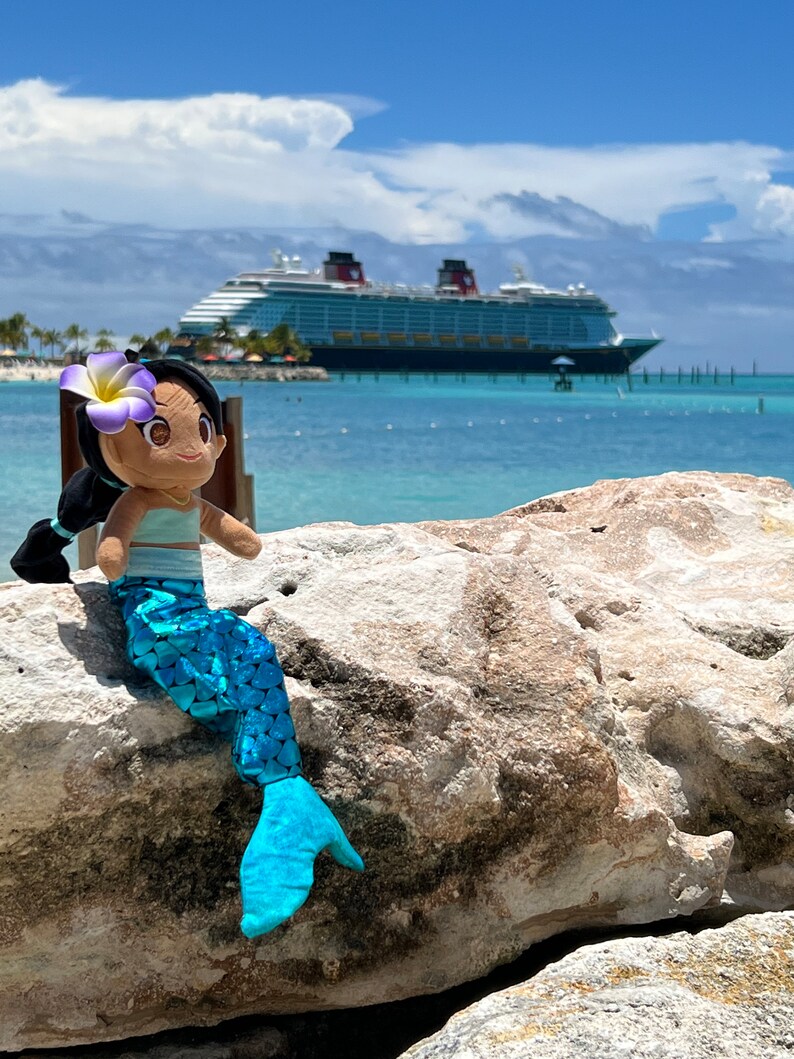 Puede incluir: Una mu&ntilde;eca de sirena de peluche con cabello casta&ntilde;o, una flor morada y una cola azul se sienta en una roca con un gran crucero en el fondo.