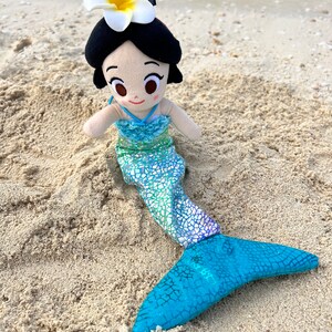 Puede incluir: Una mu&ntilde;eca de sirena de peluche con una flor blanca en la cabeza, que lleva una cola con estampado verde y azul. La mu&ntilde;eca est&aacute; sentada en la arena.